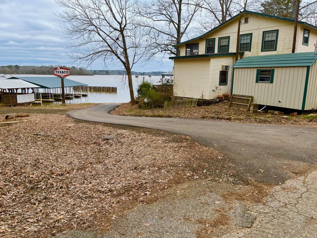 A winding road leads to a waterfront with a two-story beige house on the right and a dock with a Texaco sign on the left, surrounded by leafless trees under a cloudy sky.