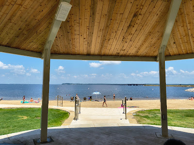 View from under a wooden pavilion looking out onto a sandy beach with people sunbathing and swimming in the water. The sky is blue with scattered clouds, and there is a small island in the distance.