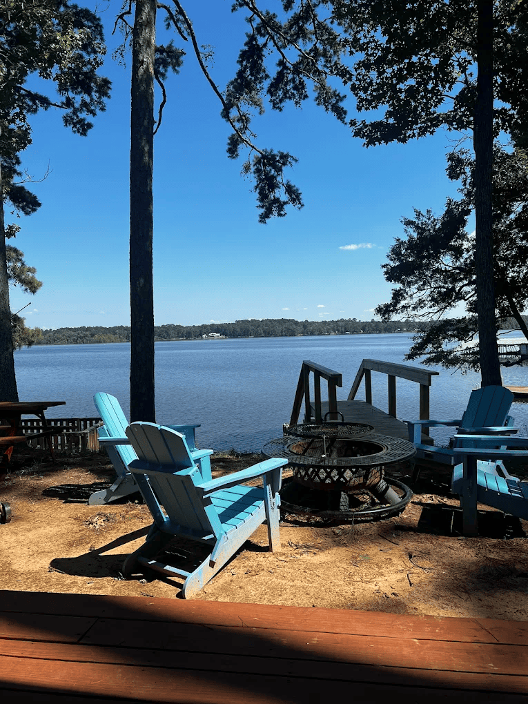 Four blue Adirondack chairs surround a fire pit on a lakeside patio, with a wooden dock and trees framing a view of the water under a clear blue sky.