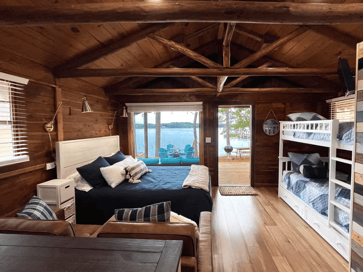 A rustic cabin bedroom with a double bed, bunk beds, wood walls and ceiling, and a view of a lake and deck through glass doors.