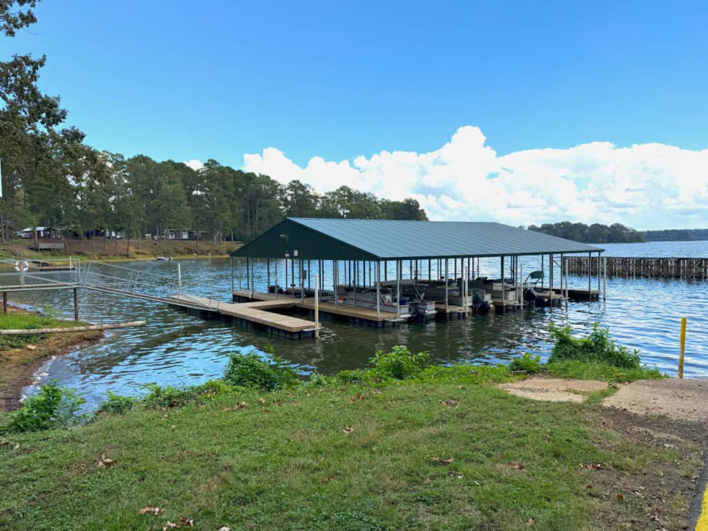 A covered boat dock extends into a lake, surrounded by trees and grassy shoreline under a partly cloudy sky.