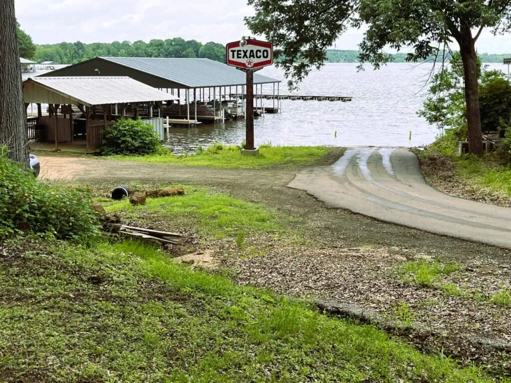 A small paved road leads to a dock and boathouse by a lake, with a vintage Texaco sign on a pole near the shoreline. Trees and grass surround the area.