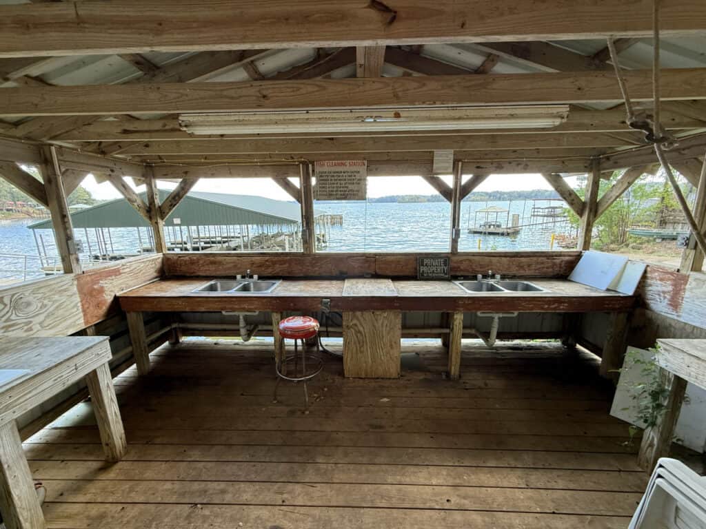 Wooden outdoor cleaning station with three sinks, a red stool, and signs, overlooking a lake with boat docks visible in the background.