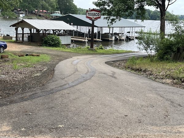 A paved road slopes down toward a lakeside marina with covered boat docks. An old Texaco sign stands near the waterfront, surrounded by trees and wooden structures. A paved road slopes down toward a lakeside marina with covered boat docks. An old Texaco sign stands near the waterfront, surrounded by trees and wooden structures.