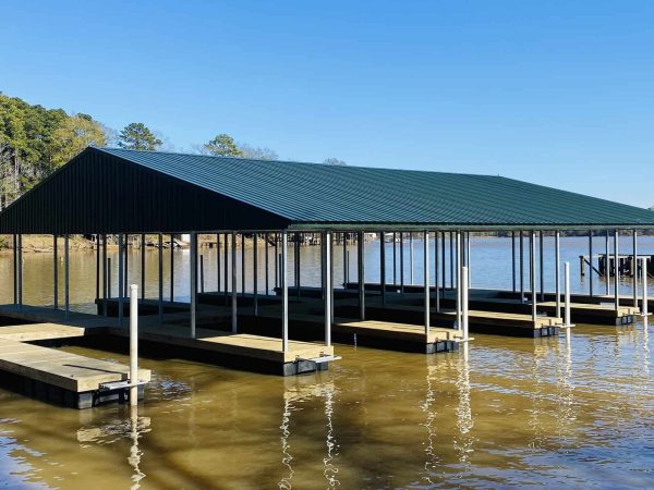 A covered boat dock with multiple slips extends over calm lake water under a clear blue sky, surrounded by trees on the shoreline.