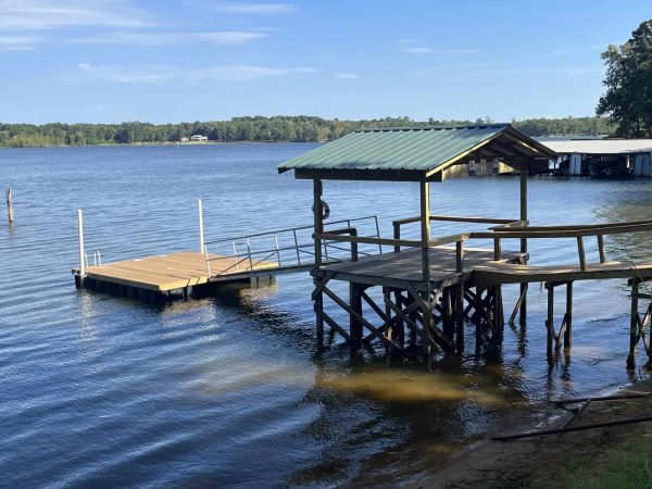 A wooden dock with a covered section extends over calm lake water, surrounded by trees and a clear sky in the background.